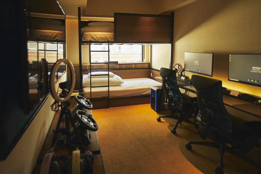 A hotel room with a loft bed, racing wheels, and a dual-monitor gaming desk