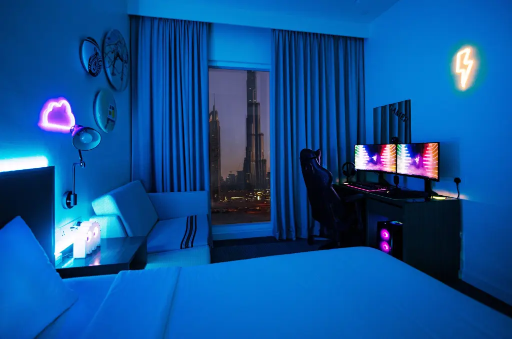 A hotel room with a neon-lit gaming PC setup and a city skyline view